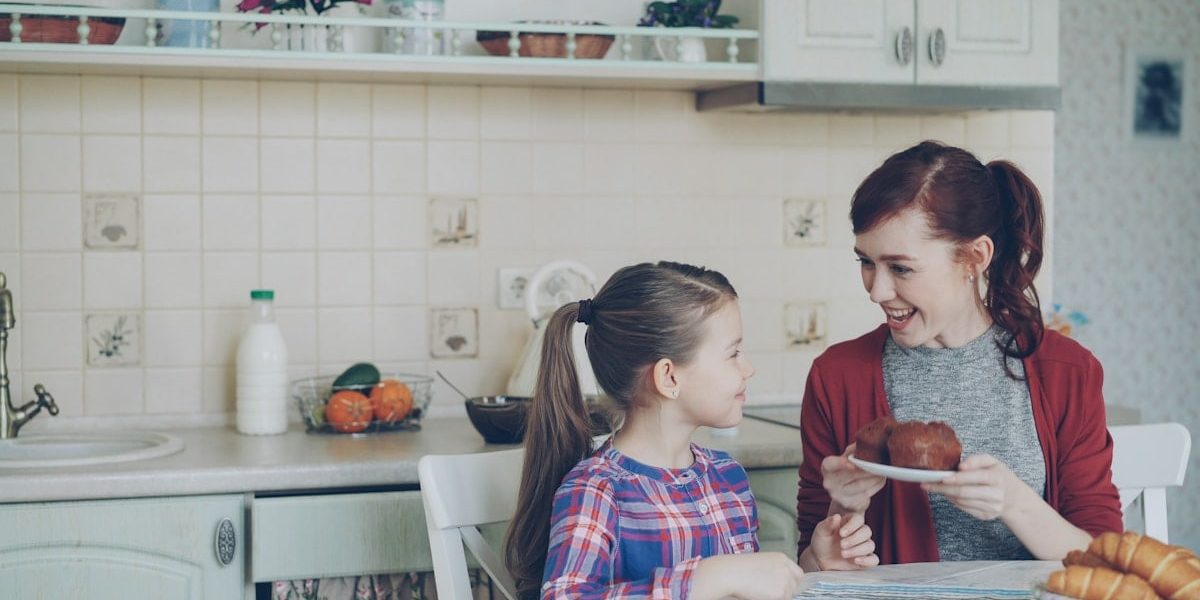 Young happy mother bringing tasty muffins to her cute daughter sitting at the table in kitchen waiting for breakfast at home