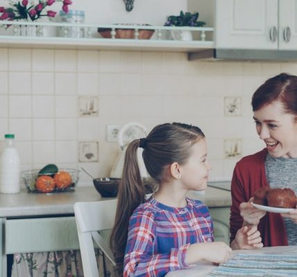 Young happy mother bringing tasty muffins to her cute daughter sitting at the table in kitchen waiting for breakfast at home